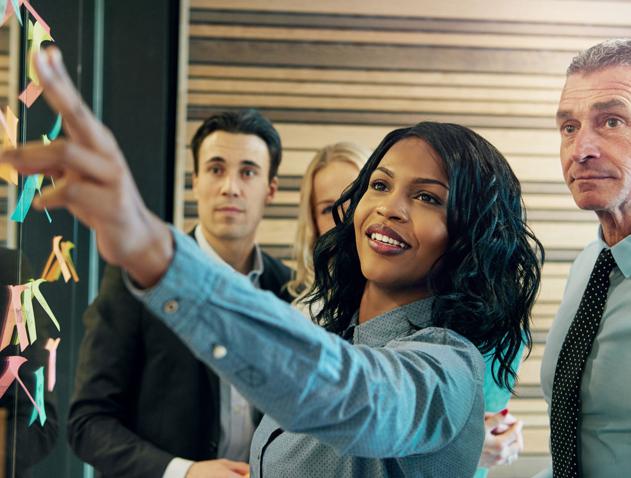 A diverse group of professionals during a brainstorming session talk and contemplate colorful sticky notes placed on a wall, as the woman in the center of the frame wearing a blue patterned shirt smiles and points to a particular note.