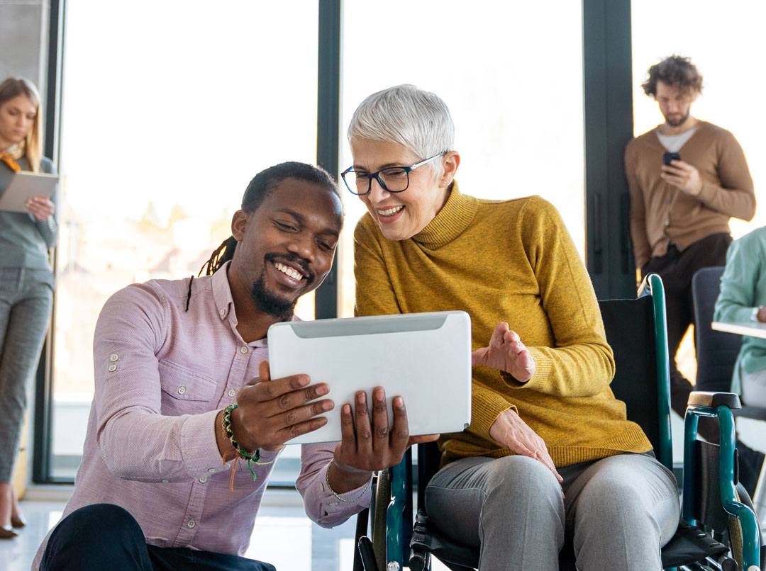 A group of people use their digital devices in a modern office setting. Center, an older woman in a wheelchair and a younger man with a pink shirt share a tablet and smile.