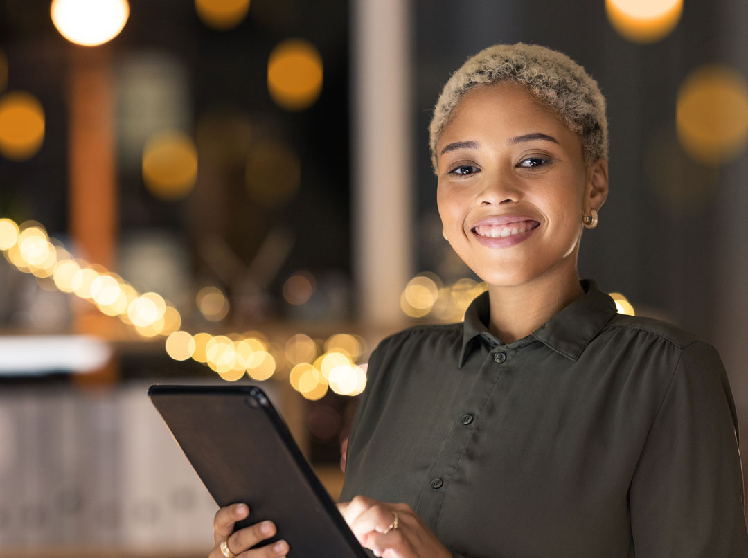 A young woman in black and tan uses a black laptop while smiling at the camera in a bulb-lit office space.